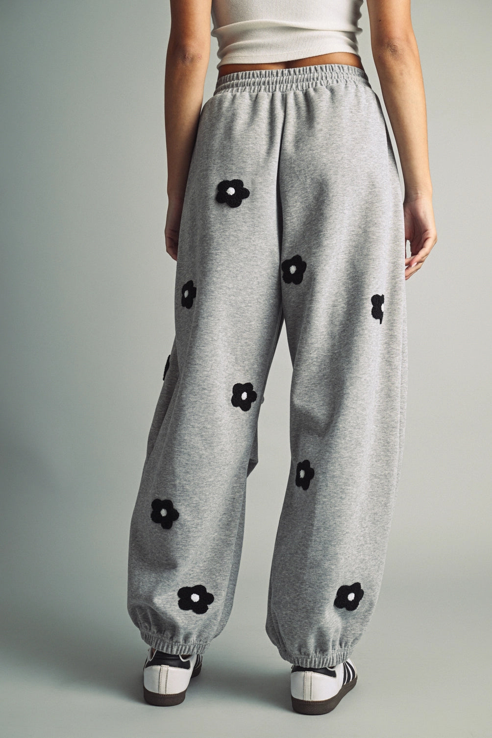 A women's heather gray wide-leg fleece sweatpants featuring an elastic drawstring waistband, relaxed barrel leg, and black chenille flower patches across the front and back.