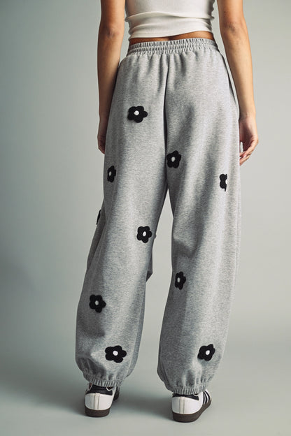 A women's heather gray wide-leg fleece sweatpants featuring an elastic drawstring waistband, relaxed barrel leg, and black chenille flower patches across the front and back.