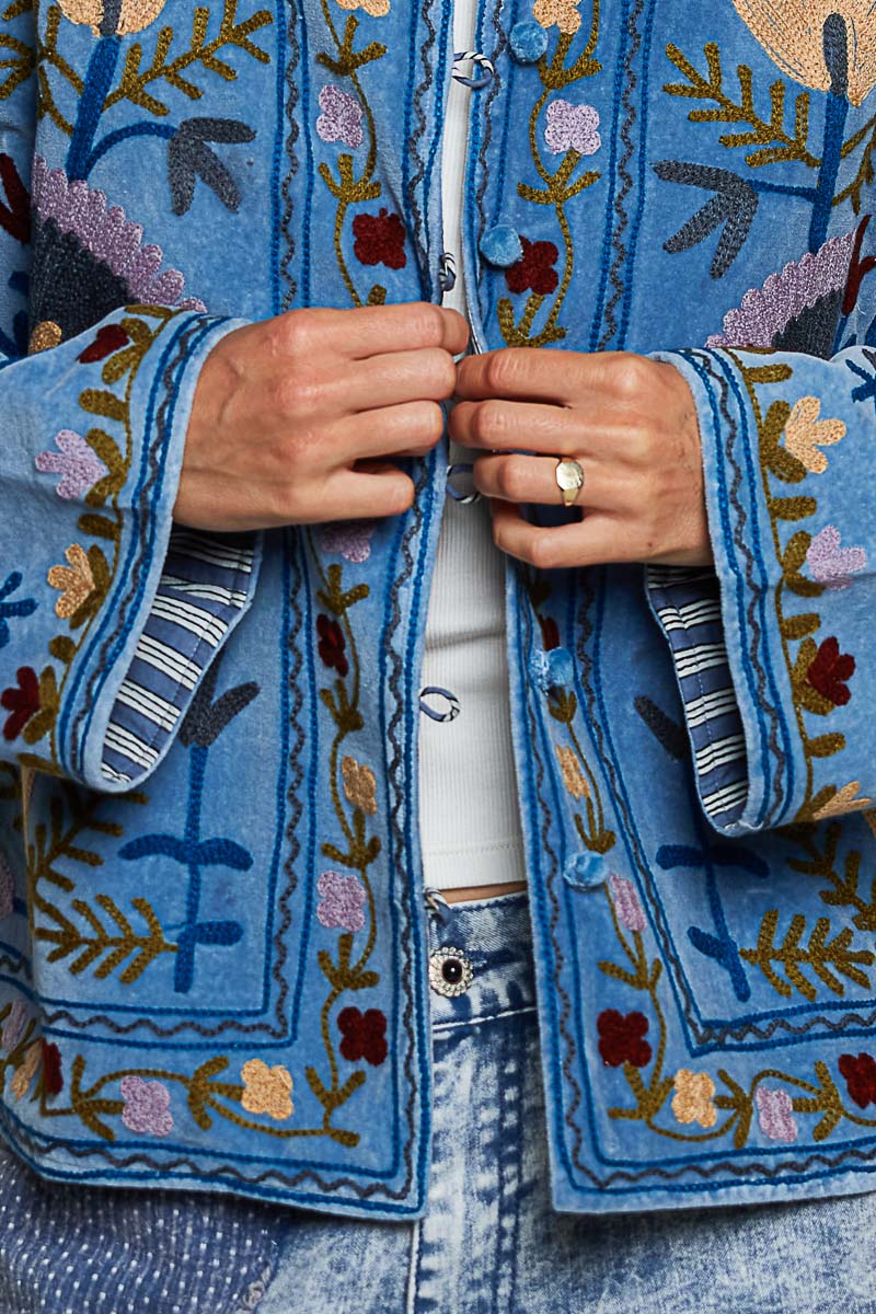 A women's dusty denim blue long-sleeve velvet jacket with allover folk floral embroidery in rust, yellow, and lavender, styled with a mandarin collar, wide cuffs, and front loop buttons.