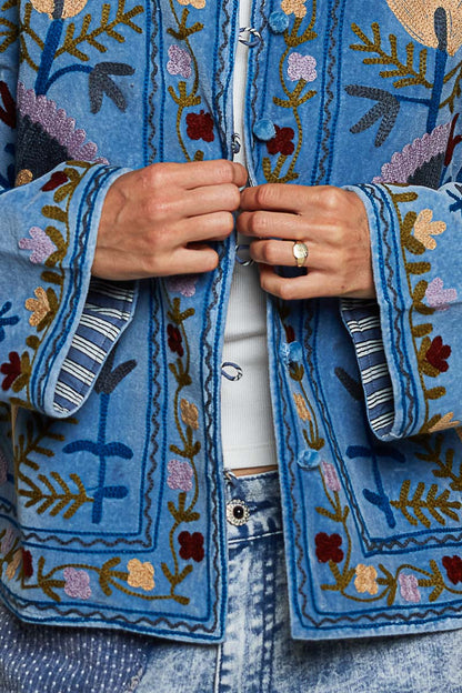 A women's dusty denim blue long-sleeve velvet jacket with allover folk floral embroidery in rust, yellow, and lavender, styled with a mandarin collar, wide cuffs, and front loop buttons.