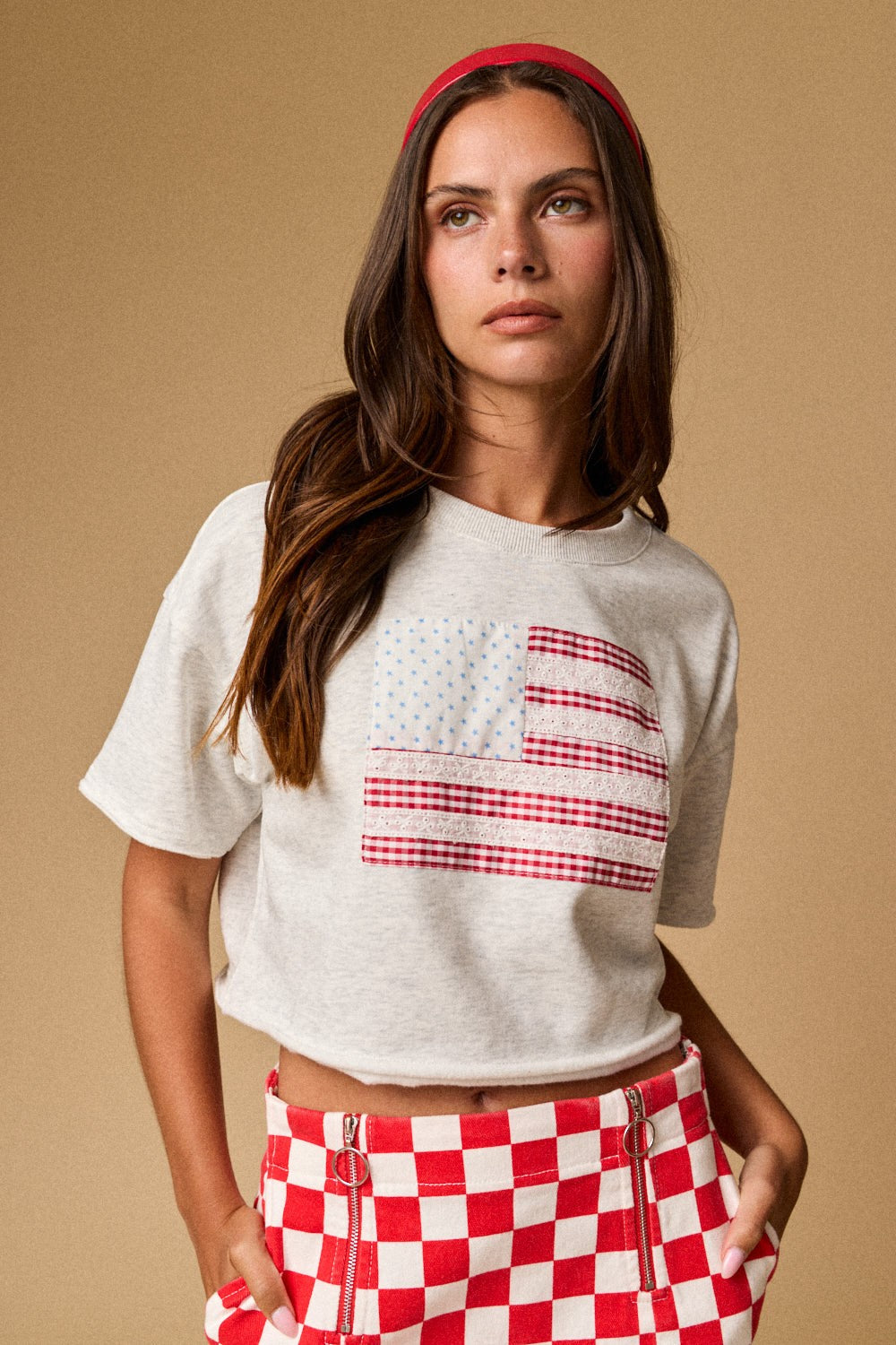 A women's light gray short-sleeve graphic t-shirt featuring a relaxed fit, a raw cut cropped hemline, and a textured patchwork American flag design made from blue star print, red gingham, and white eyelet lace fabrics.