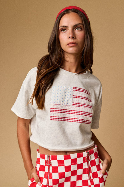 A women's light gray short-sleeve graphic t-shirt featuring a relaxed fit, a raw cut cropped hemline, and a textured patchwork American flag design made from blue star print, red gingham, and white eyelet lace fabrics.