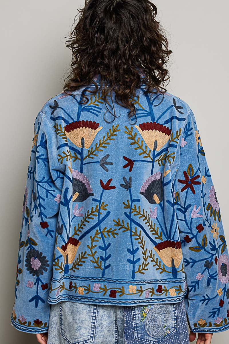 A women's dusty denim blue long-sleeve velvet jacket with allover folk floral embroidery in rust, yellow, and lavender, styled with a mandarin collar, wide cuffs, and front loop buttons.