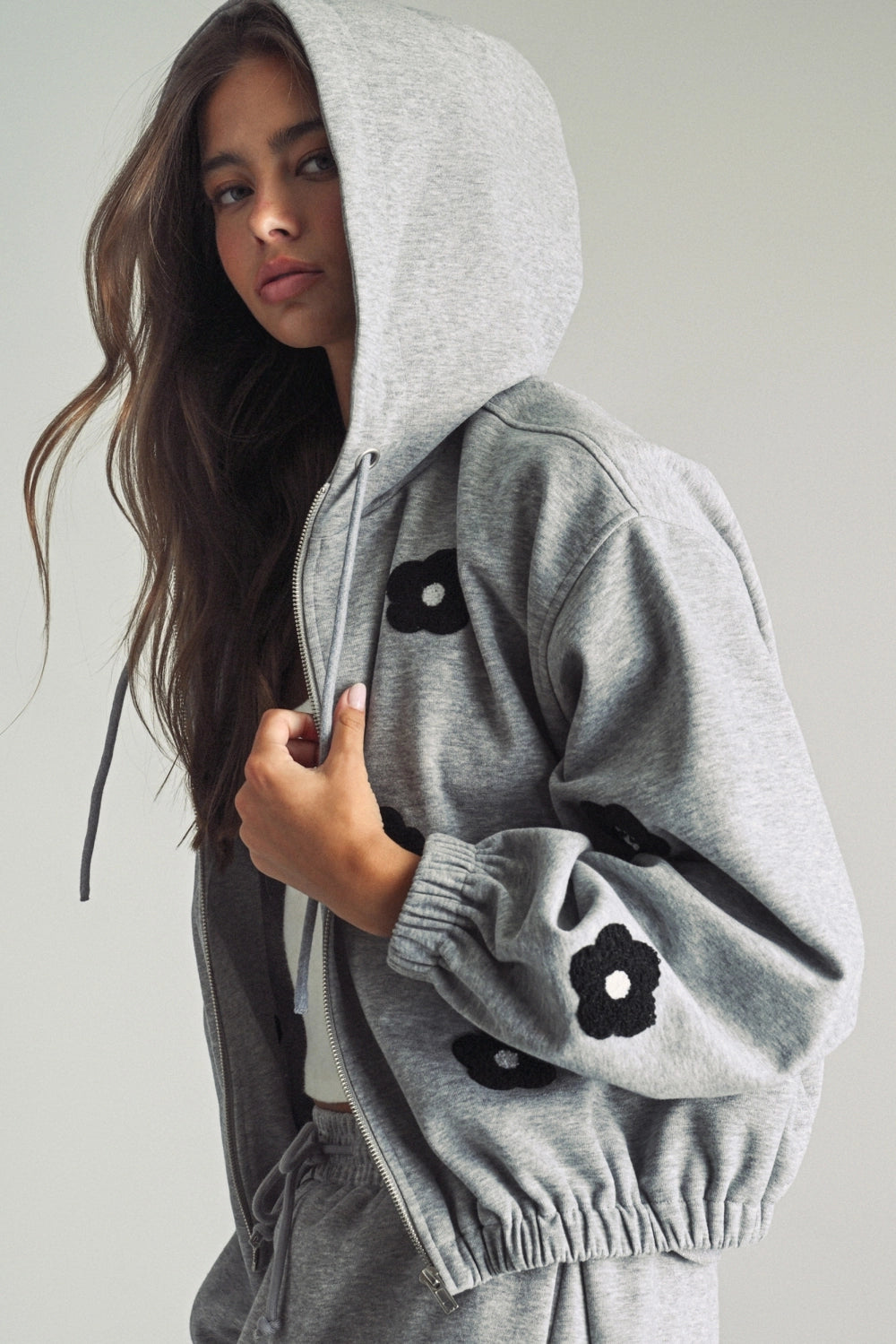 A women's heather gray full-zip hooded fleece jacket featuring dropped shoulders, ribbed hem cuffs, and black chenille flower patches on the front and sleeves.
