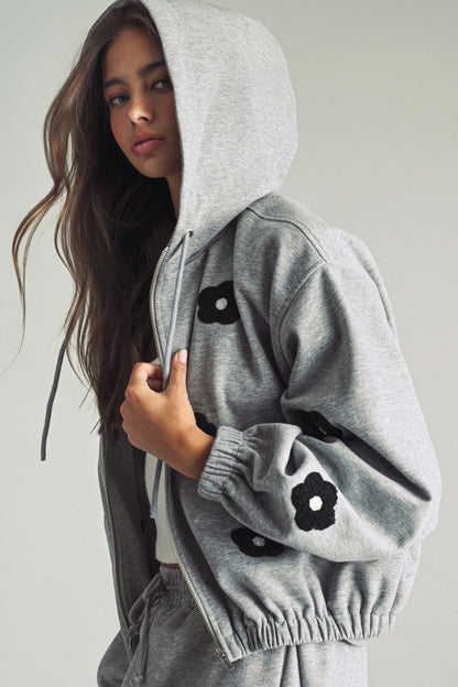 A women's heather gray full-zip hooded fleece jacket featuring dropped shoulders, ribbed hem cuffs, and black chenille flower patches on the front and sleeves.