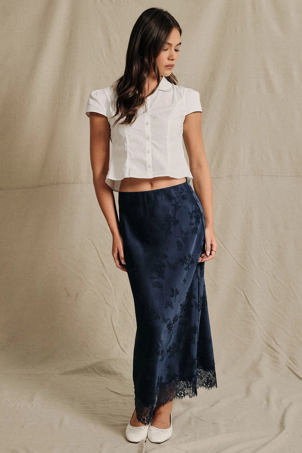 A women's navy blue satin bias-cut slip midi skirt featuring an elastic waistband and delicate scalloped lace trim along the hemline.