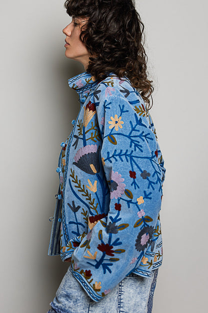A women's dusty denim blue long-sleeve velvet jacket with allover folk floral embroidery in rust, yellow, and lavender, styled with a mandarin collar, wide cuffs, and front loop buttons.