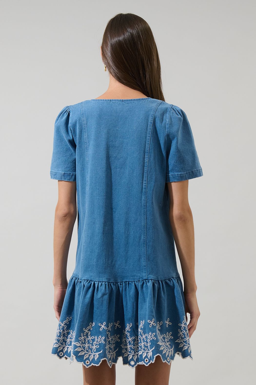 A women's medium wash blue denim short-sleeve mini dress featuring a square neckline, ruffled button placket, and cream floral embroidered scalloped hemline.