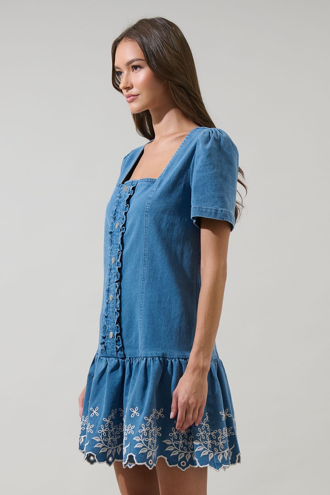 A women's medium wash blue denim short-sleeve mini dress featuring a square neckline, ruffled button placket, and cream floral embroidered scalloped hemline.