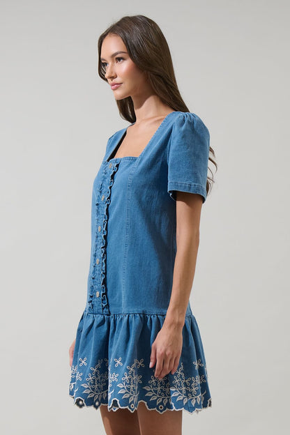 A women's medium wash blue denim short-sleeve mini dress featuring a square neckline, ruffled button placket, and cream floral embroidered scalloped hemline.