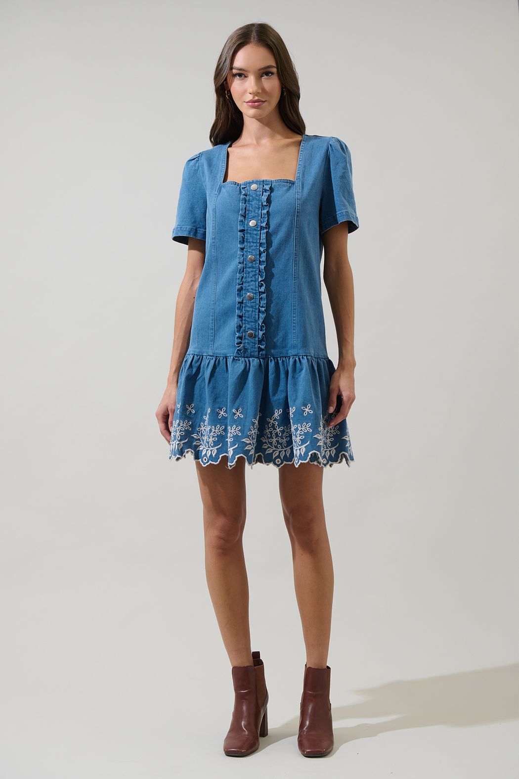 A women's medium wash blue denim short-sleeve mini dress featuring a square neckline, ruffled button placket, and cream floral embroidered scalloped hemline.