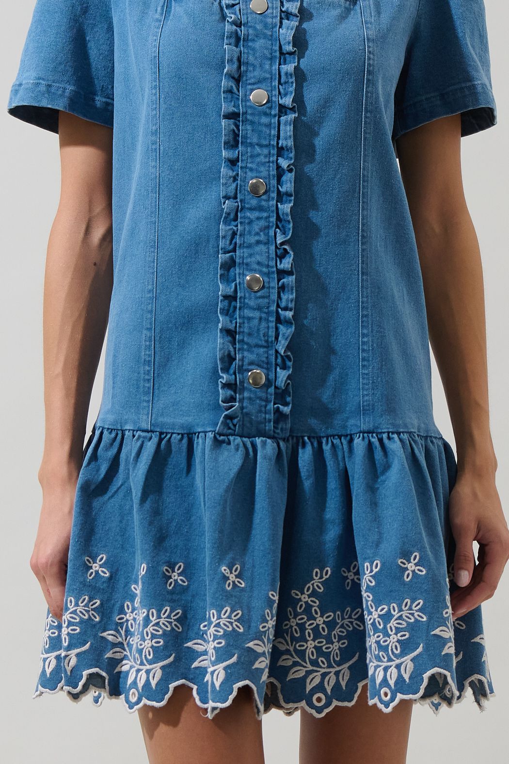 A women's medium wash blue denim short-sleeve mini dress featuring a square neckline, ruffled button placket, and cream floral embroidered scalloped hemline.